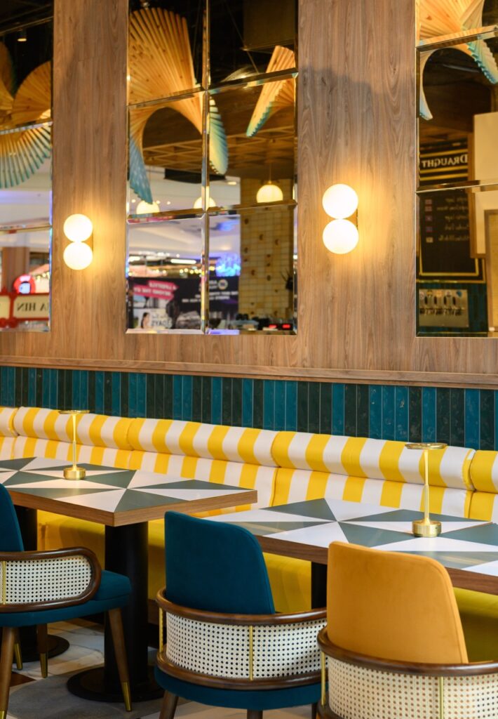 Yellow striped banquette against petrol-teal stacked tile with mirrors and globe wall lights
