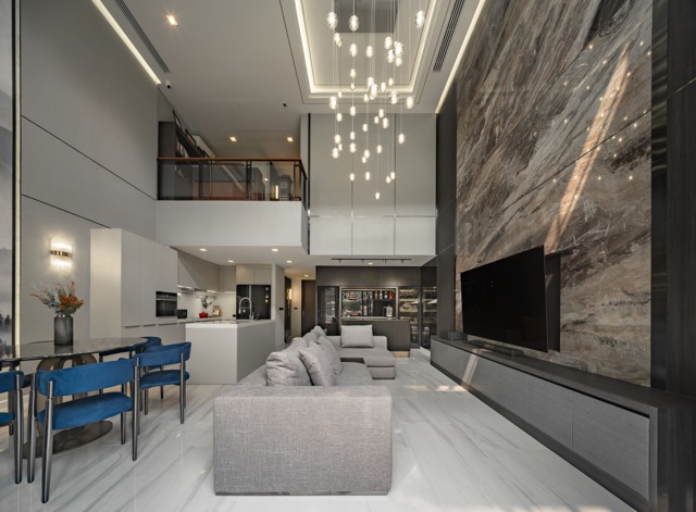 Quarter31 Bangkok — double-height living space with a full-height veined stone wall, a cascading pendant cluster through the void, mezzanine above and blue dining chairs at the open kitchen
