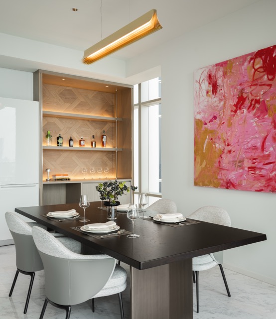 Dining room with dark wood table, backlit bar cabinet, brass pendant and pink abstract painting in a Bangkok luxury residence