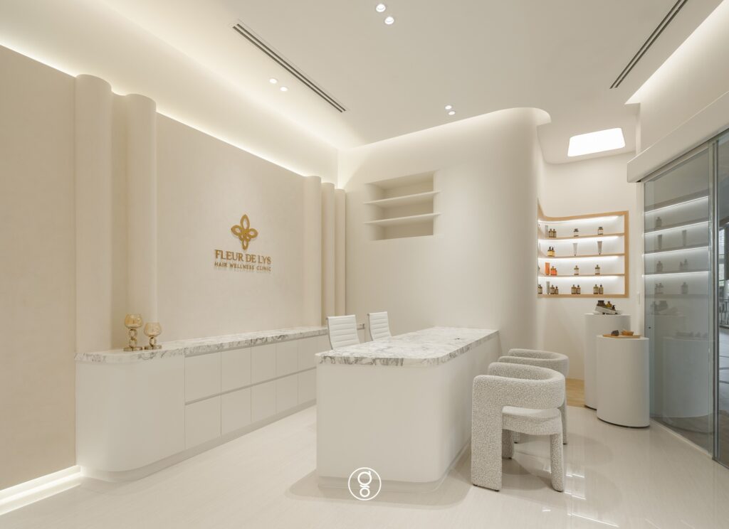 Fleur De Lys Hair Wellness Clinic, K Village — white reception with marble consultation counter, sculpted plaster walls and backlit product niches, by Gratitude Design