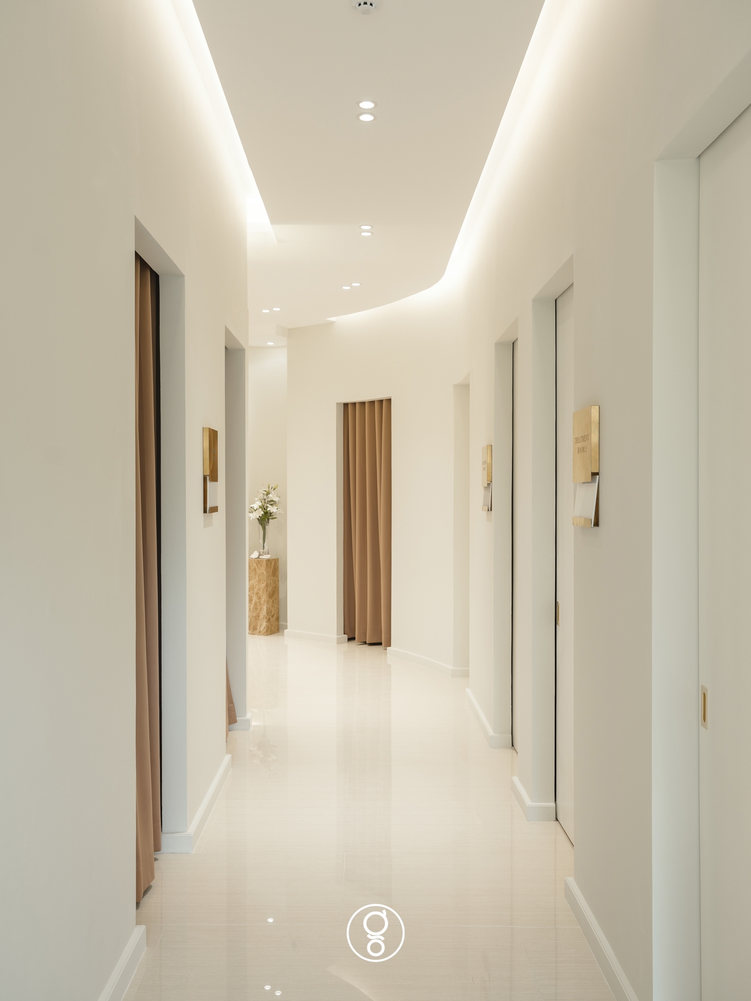 Clinic corridor in cream with brass room plates, continuous cove lighting and a curtained opening at the far end