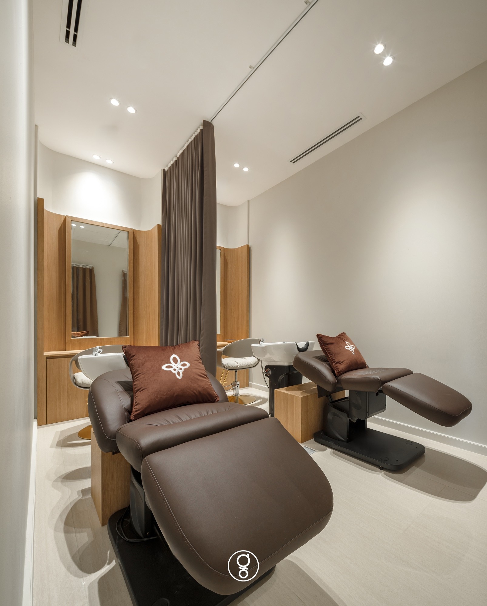Scalp treatment and wash bay with two brown leather reclining wash chairs, oak mirror units and branded cushions behind a grey curtain