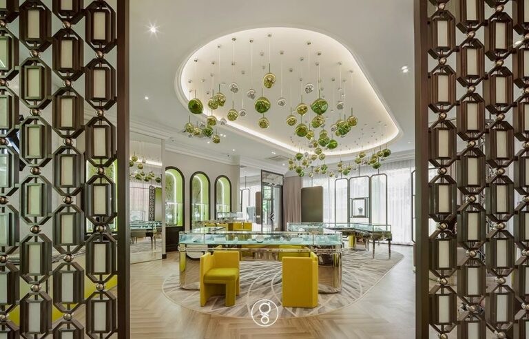 Scintillion Surawongse jewellery showroom — glass display tables beneath a sculpted ceiling of green glass pendants, framed by bronze lattice screens, by Gratitude Design