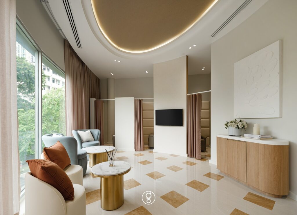 Clinic waiting lounge with a gold illuminated ceiling disc, blue and cream tub chairs, brass and marble side tables and a travertine chequer floor