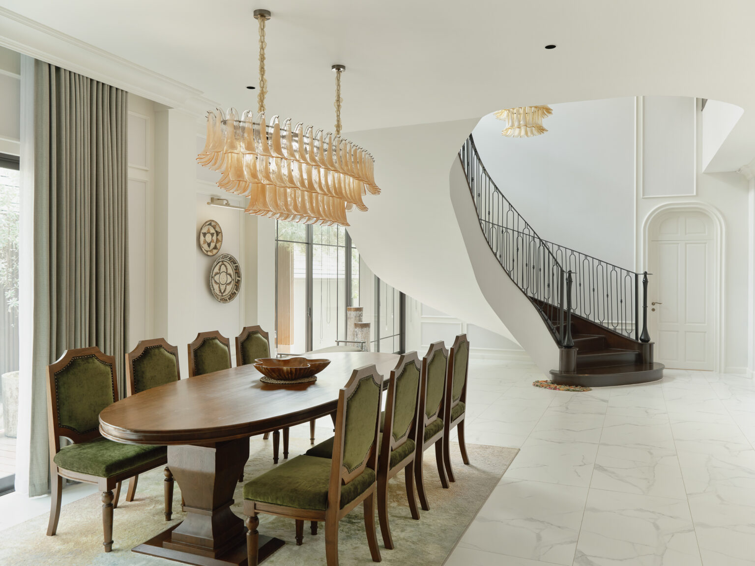 Grand Bangkok Boulevard dining room — oval walnut table with olive velvet chairs under a tiered amber glass chandelier, curved staircase beyond, interior design by Gratitude Design