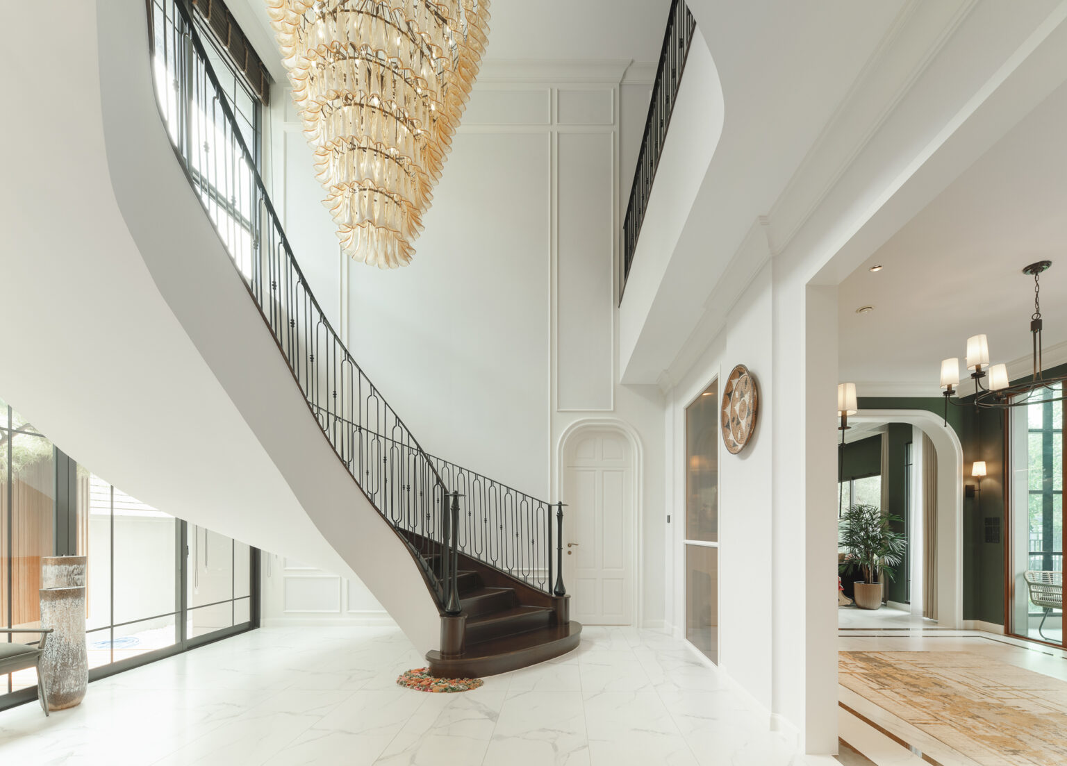 Grand Bangkok Boulevard double-height entrance hall — sculpted white staircase with a black wrought-iron balustrade beneath a tiered amber glass chandelier, marble floor, interior design by Gratitude Design