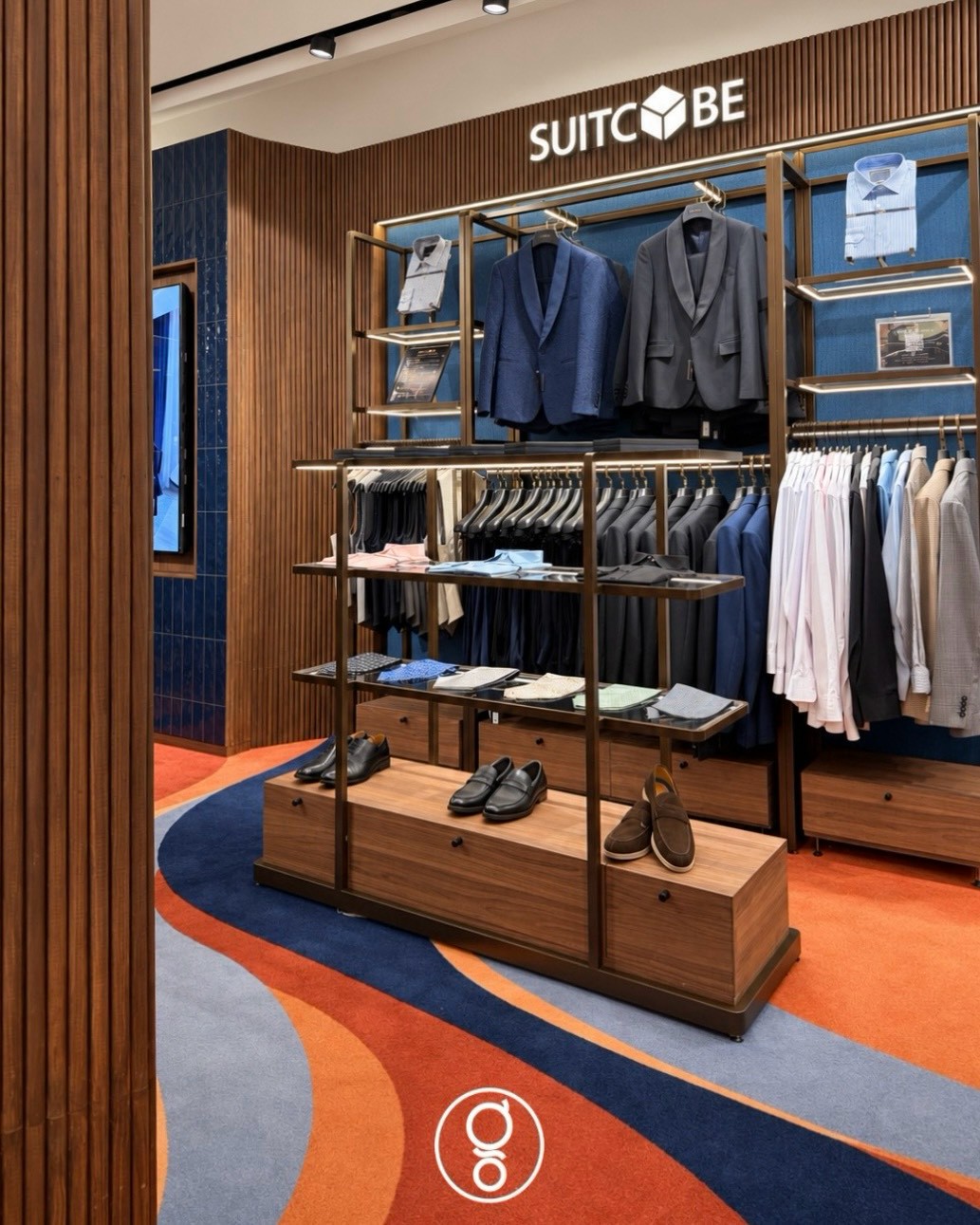 Suitcube brand wall in fluted walnut with halo-lit lettering, brass-framed shelving of suits and shirts on a deep-blue fabric backing, shoe plinths on the wave carpet