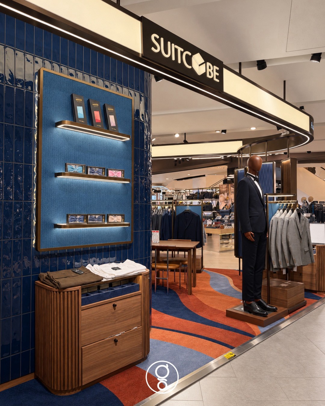 Blue glazed tile wall with a fabric-backed accessory panel of pocket squares and bow ties on lit brass shelves, Suitcube fascia and a tuxedo mannequin beyond
