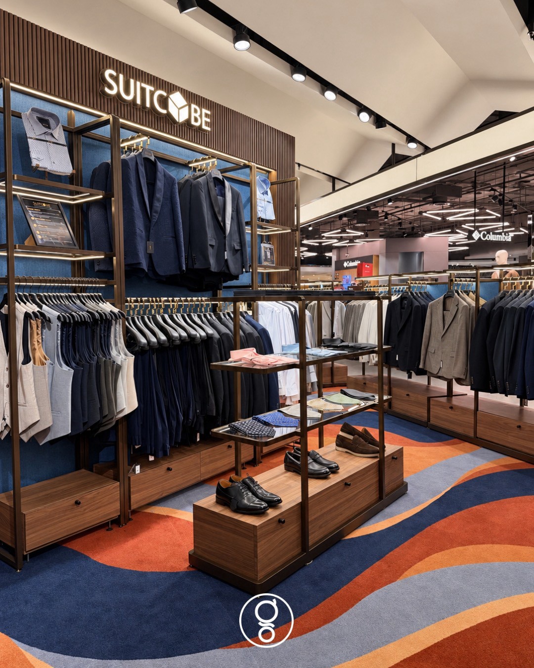 Brass-framed suit wall with waistcoats and jackets, a central accessory island with pocket squares and shoes, and the department-store floor beyond