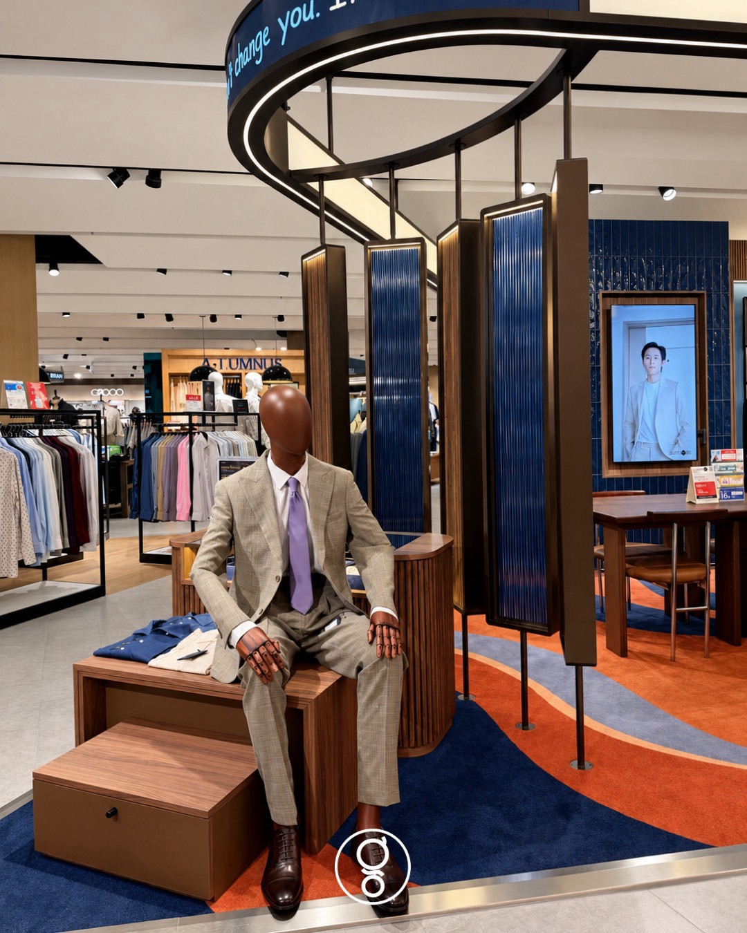 Seated mannequin in a pale check suit on a walnut plinth beneath the oval canopy, hanging fluted walnut and blue glass screens and a framed digital screen behind