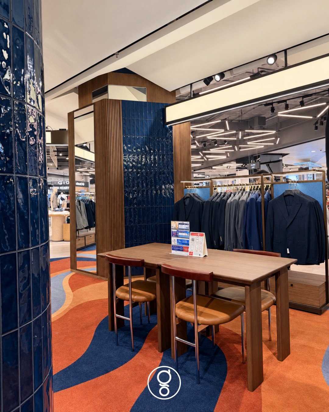 Consultation table in walnut with leather-seated chairs beside a deep-blue glazed tile column and a full-height mirror, suit rail behind
