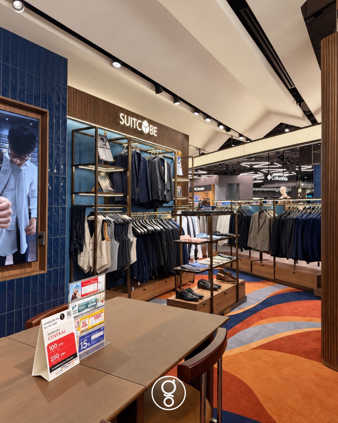 Brand wall and suit rails seen from the consultation table, a framed digital screen set into the blue tile wall, wave carpet in navy and terracotta