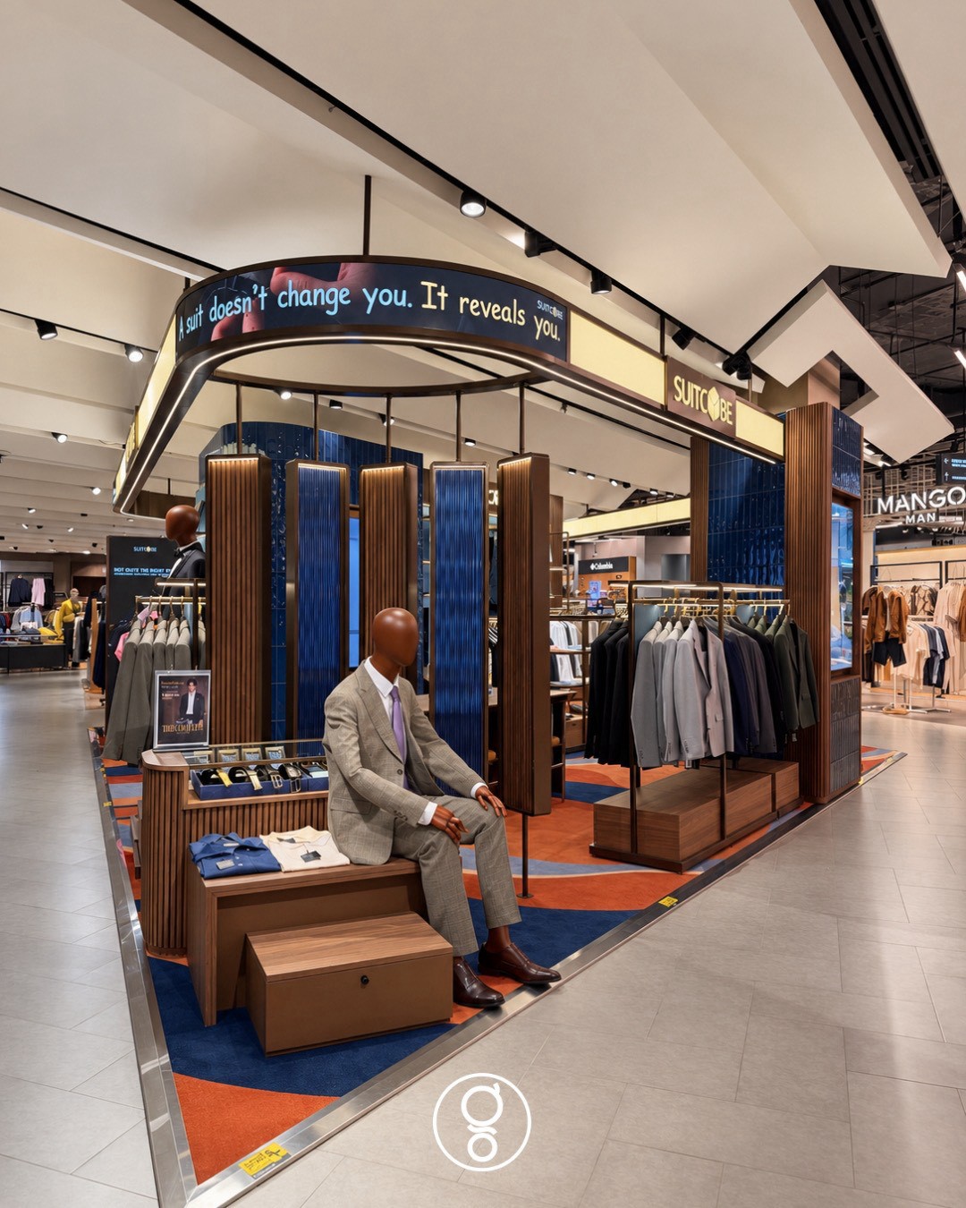 Suitcube Central World menswear store interior design in Bangkok, shopfront on the department-store floor with an oval brass canopy, fluted walnut piers and a wave-patterned carpet
