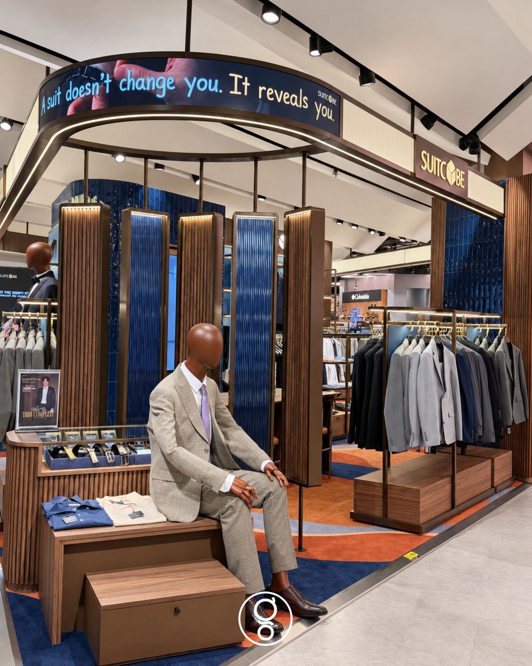 Shopfront from the aisle — seated mannequin on a walnut plinth, accessory counter, brass suit rail and the Suitcube sign on the canopy