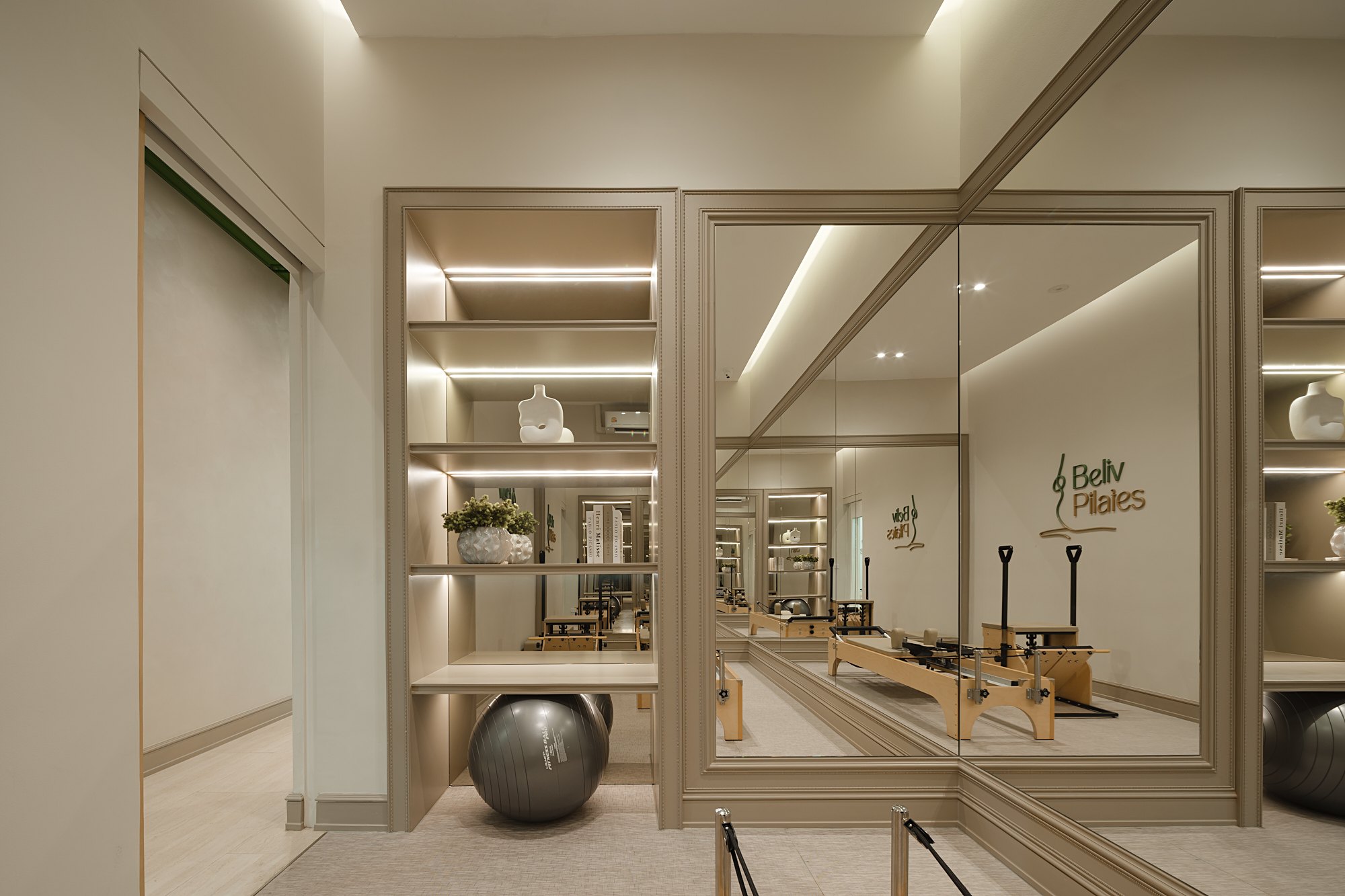 Beliv Pilates — ชั้นบิลท์อินซ่อนไฟกับของตกแต่งและลูกบอลออกกำลังกาย ข้างแผงกระจกเงาที่สะท้อนห้องรีฟอร์มเมอร์