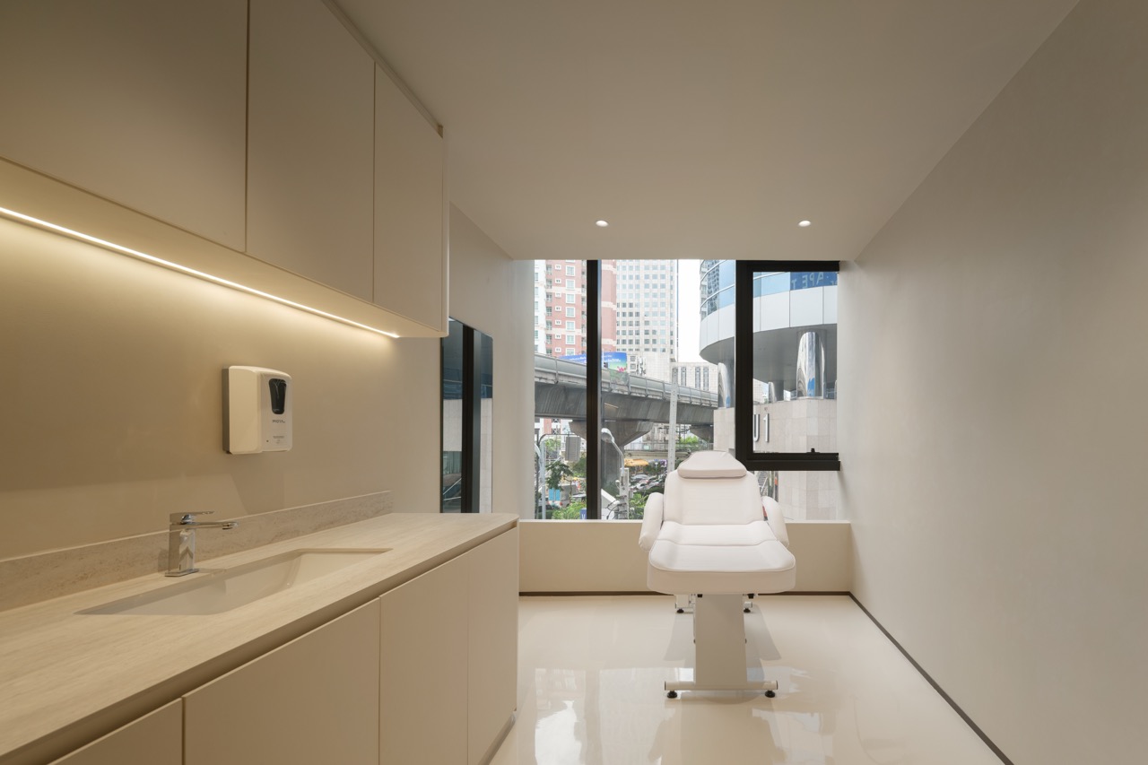 ห้องทำหัตถการ Hairtran Clinic — เก้าอี้ทำหัตถการสีขาวริมหน้าต่าง เคาน์เตอร์อ่างท็อปหิน และตู้บานเรียบพร้อมไฟใต้ตู้