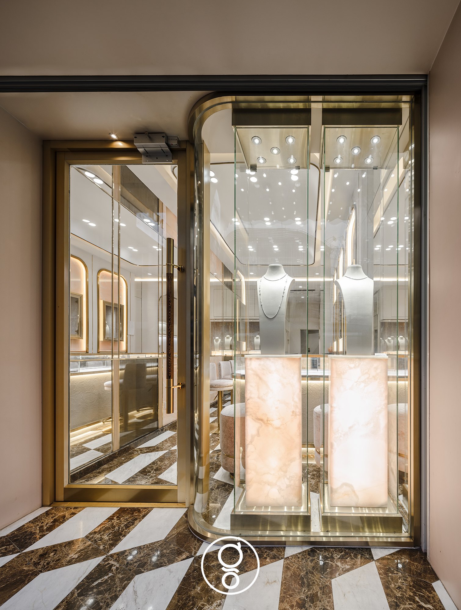 Nopparat Jewelry storefront — a brass-framed glass door beside a curved glass showcase with necklace busts on backlit onyx plinths, on a geometric marble floor