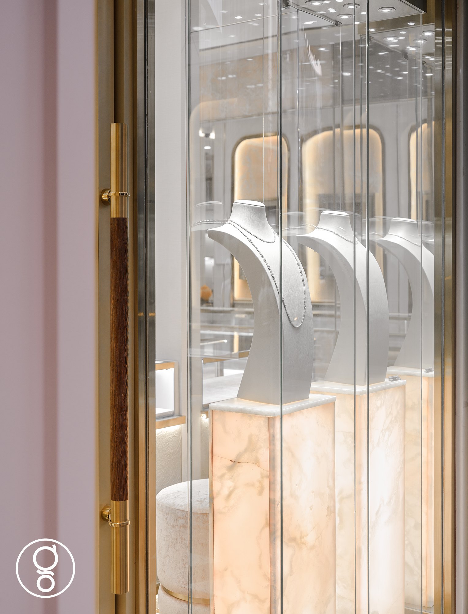 Window showcase at Nopparat Jewelry — necklace busts on backlit onyx plinths behind glass, beside a brass and timber door handle