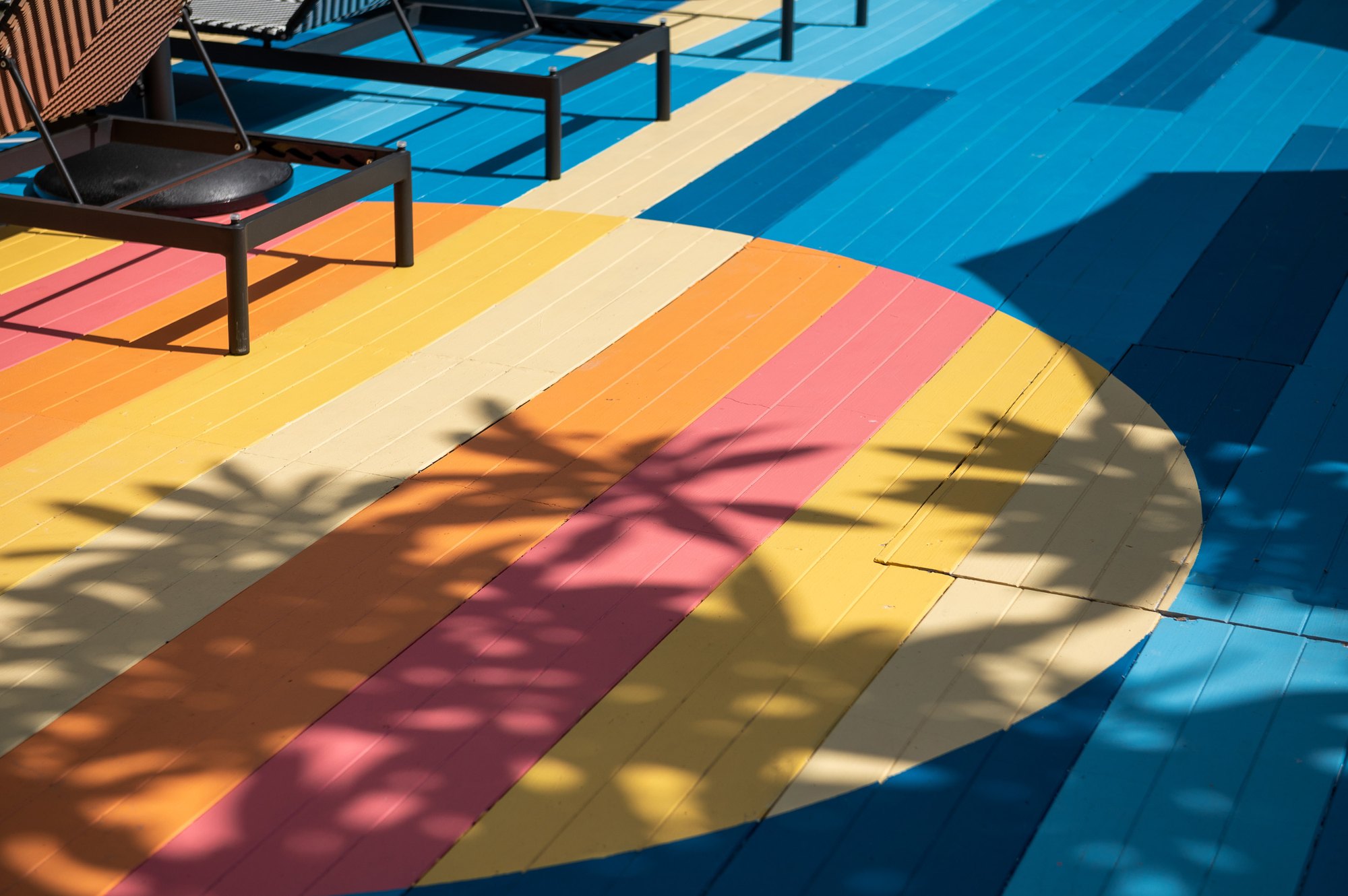 Painted pool deck at Sala Huahin — a circle of orange, pink and yellow stripes on blue boards, with palm shadows and lounger frames