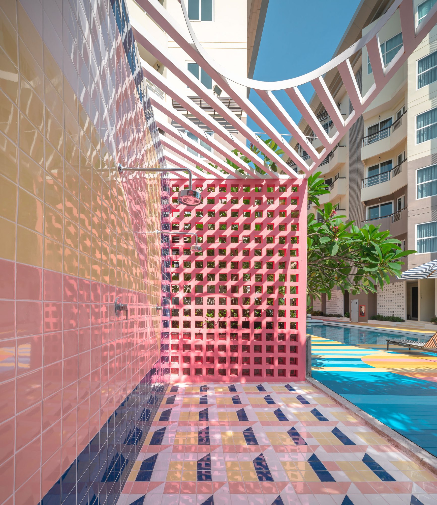 Sala Huahin outdoor shower — pink and yellow glazed tiles, a coral breeze-block screen, a curved white pergola and a geometric tiled floor beside the pool