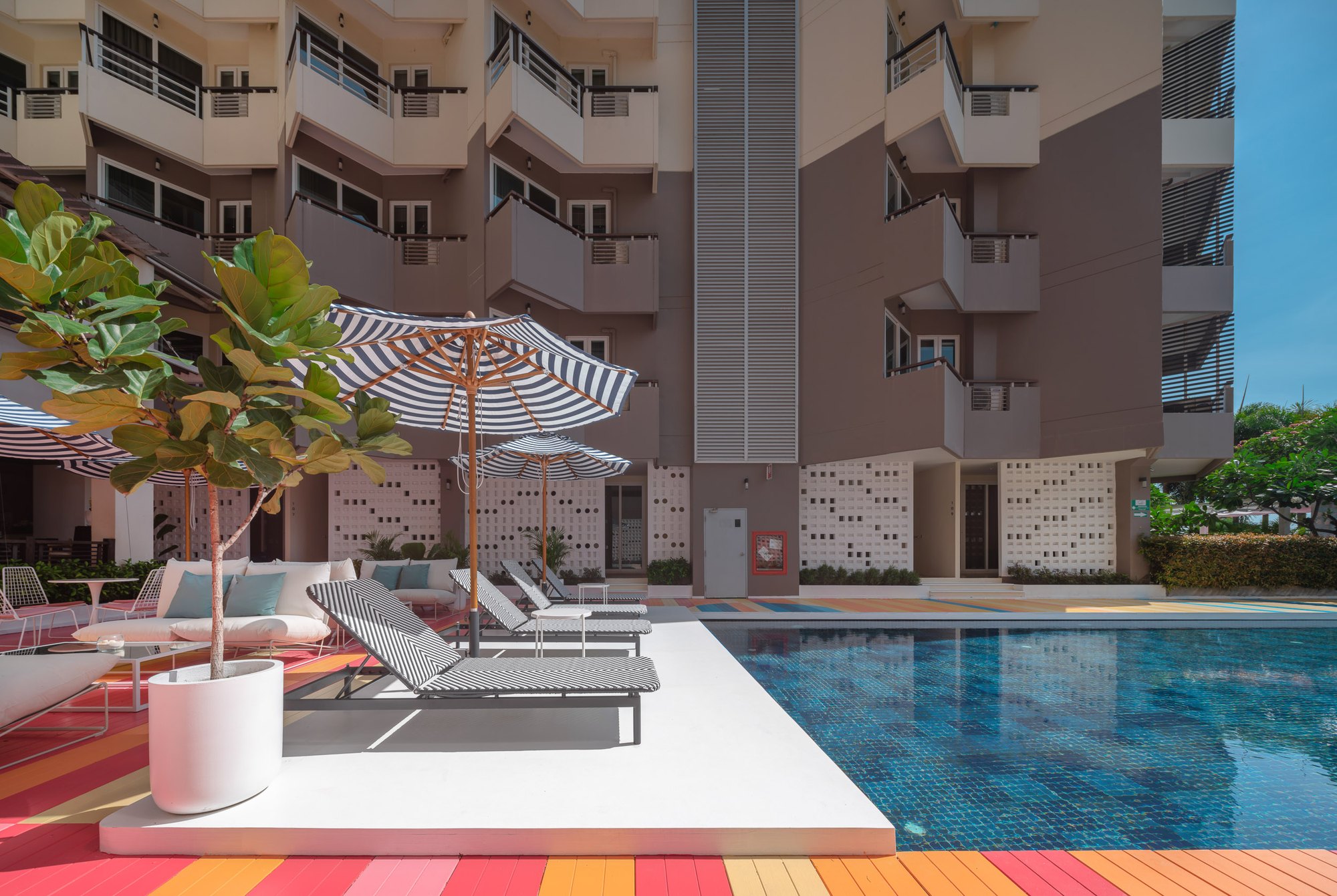 Sala Huahin — loungers and parasols on a white platform at the pool edge, white sofas with blue cushions and a coral striped deck, breeze-block screens beyond